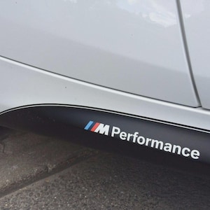 Op de afbeelding: Een wit zijpaneel van een auto met een matzwarte dorpelsticker. De sticker bevat het M Performance-logo in witte letters, vergezeld van een grafisch element bestaande uit drie schuine strepen in lichtblauw, donkerblauw en rood.