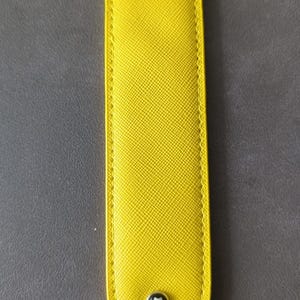 K&ouml;nnte beinhalten: Ein leuchtend gelbes, l&auml;ngliches und abgerundetes Stifteetui aus Leder mit einer strukturierten Kreuzschraffur-Oberfl&auml;che und farblich abgestimmten N&auml;hten an den R&auml;ndern. Ein kleines, rundes, silberfarbenes Emblem mit einem wei&szlig;en sechsstrahligen Stern auf schwarzem Grund ist unten mittig angebracht.