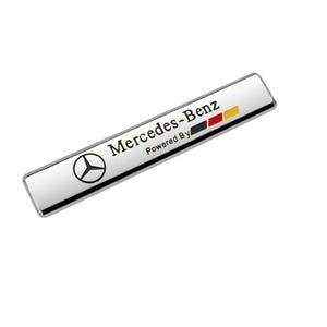 Puede incluir: Un emblema rectangular de metal plateado que presenta el logotipo negro de Mercedes-Benz a la izquierda. El texto dice Mercedes-Benz Powered By, seguido de tres pequeñas barras horizontales en colores negro, rojo y amarillo a la derecha.