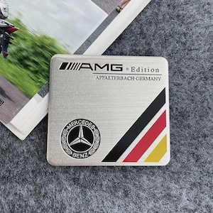 May include: A rectangular brushed silver metal emblem featuring the black AMG Edition logo and the text AFFALTERBACH GERMANY. It includes a circular black and silver Mercedes-Benz logo and three diagonal stripes in black, red, and yellow colors.