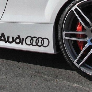 May include: A white car side panel featuring a black Audi logo decal with the brand name and four rings. The vehicle has a large silver multi-spoke alloy wheel with a visible red brake caliper positioned behind the spokes on a dark asphalt surface.