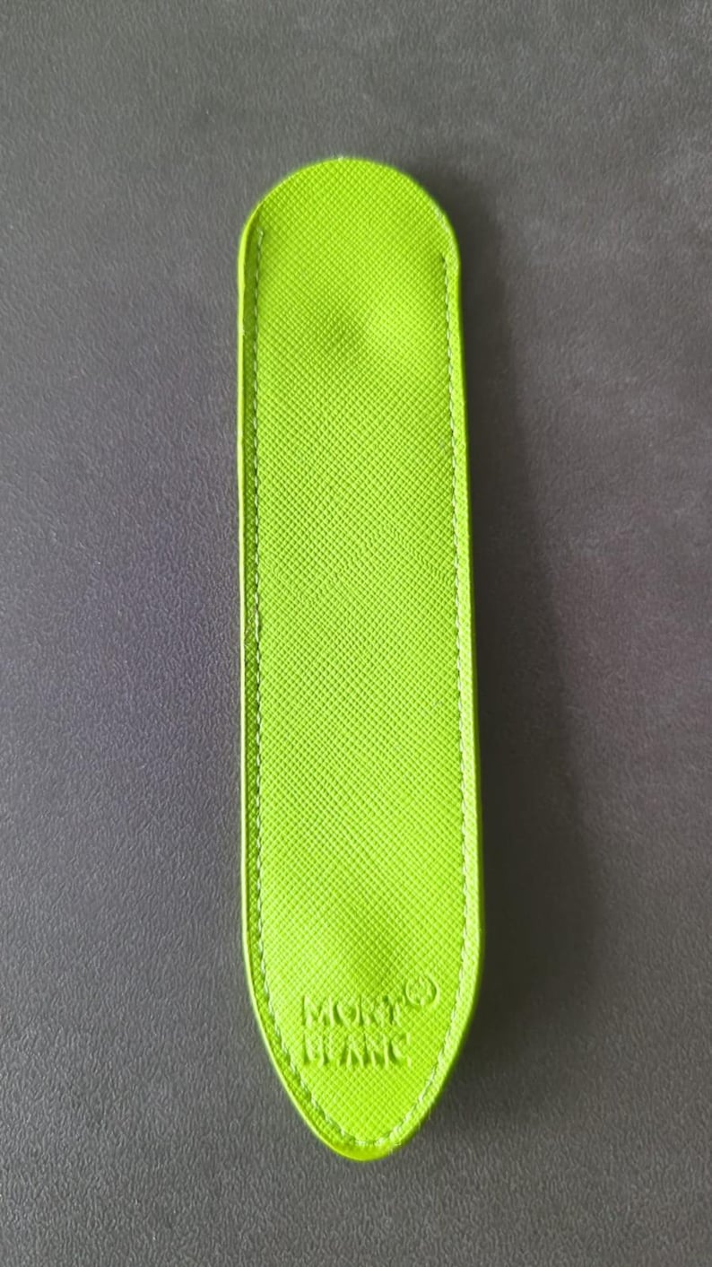 May include: A lime green pen pouch with a textured surface and rounded ends. The accessory features visible stitching along the edges and the embossed text MONTBLANC with a small circular logo symbol located near the bottom of the front side.