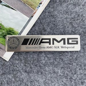 Puede incluir: Un emblema rectangular de metal plateado que presenta el logotipo de Mercedes-Benz a la izquierda, seguido del texto negro AMG y la inscripción Mercedes-Benz AMG SLK Webspecial en una fuente más pequeña en el borde inferior.