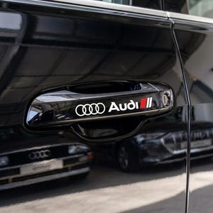 Op de afbeelding: Een glanzende zwarte autodeurklink met het witte Audi-logo en een rood en zwart rechthoekig grafisch accent. De klink is geïntegreerd in het gladde, reflecterende zwarte carrosseriepaneel van het voertuig en heeft een strak ontwerp.