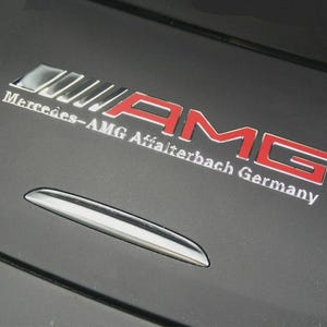 Könnte beinhalten: Ein metallisches Emblem mit dem roten und silbernen Schriftzug AMG und dem darunter liegenden silbernen Text Mercedes-AMG Affalterbach Germany auf einer mattschwarzen Oberfläche mit einem geschwungenen silbernen Akzent unter dem Schriftzug.