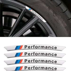 Op de afbeelding: Vier gebogen witte stickers voor autovelgen. Elke sticker heeft aan de linkerkant een grafisch motief met schuine blauwe, lichtblauwe en rode strepen, gevolgd door het woord Performance in zwarte letters. De stickers zijn aangebracht op een donkergrijze velg.