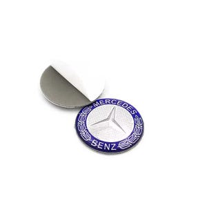 May include: A circular adhesive emblem featuring the Mercedes-Benz logo in silver on a textured background, surrounded by a blue border with the text MERCEDES BENZ and a laurel wreath design. A separate circular adhesive backing is visible nearby.