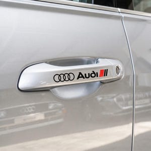 Op de afbeelding: Een zilveren autodeurklink met het zwarte Audi-logo en de tekst met een rode accentstreep. De klink is geïntegreerd in het gladde, zilverkleurige carrosseriepaneel van het voertuig, met een strak ontwerp en een sleutelgat aan de rechterkant.