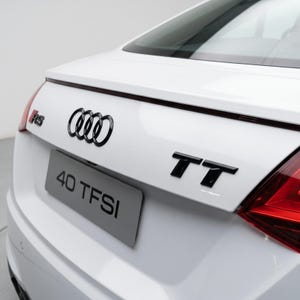 Op de afbeelding: Het achteraanzicht van een wit Audi TT-voertuig met zwarte emblemen, waaronder het logo met vier ringen, het TT-modelbadge en een RS-badge. Een kentekenplaatgedeelte toont de tekst 40 TFSI in zwarte letters op een grijze achtergrond.