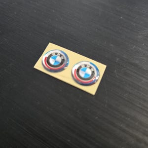 Może przedstawiać: Dwie okrągłe naklejki z logo BMW o błyszczącym, wypukłym wykończeniu na prostokątnym beżowym arkuszu. Logo przedstawia kultowy niebiesko-biały wzór ćwiartek otoczony czarnym pierścieniem z białym napisem BMW i czerwonymi akcentami.