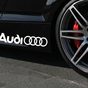 May include: A white Audi logo decal applied to the side of a black car body panel. The decal features the word Audi in a bold white font followed by the four interlocking rings logo. A silver multi-spoke alloy wheel with a red brake caliper is visible.