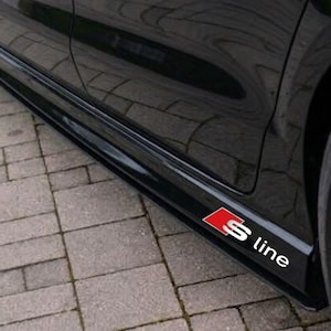 Peut inclure: Un bas de caisse de voiture noir arborant un emblème S line rouge et blanc. Le panneau noir brillant longe le bord inférieur de la portière du véhicule, positionné au-dessus du bas de caisse, avec une roue noire visible sur la droite.
