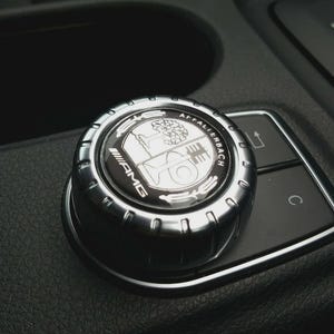 Puede incluir: Un mando de control circular de metal plateado con el logotipo de AMG y el escudo de Affalterbach en el centro. El mando está montado sobre un panel de consola negro texturizado con un pequeño botón marcado con una C y un icono de flecha.
