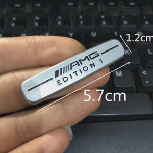 Puede incluir: Un emblema metálico rectangular de color plateado con el texto negro AMG y EDITION 1 junto a una línea horizontal. El emblema mide 5,7 cm de largo y 1,2 cm de alto, tal como indica el texto impreso directamente sobre la imagen.