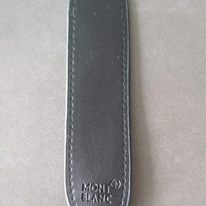 May include: A slim, elongated black leather pen pouch with rounded ends and visible stitching along the edges. The surface features a textured grain and the embossed Montblanc logo with its signature star emblem at the bottom center of the item.