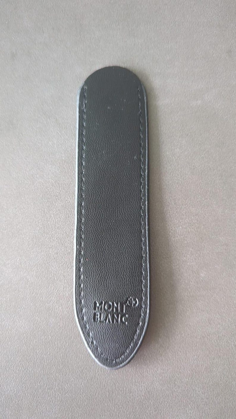 May include: A slim, elongated black leather pen pouch with rounded ends and visible stitching along the edges. The surface features a textured grain and the embossed Montblanc logo with its signature star emblem at the bottom center of the item.