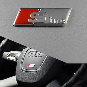 May include: A metallic S-Line emblem featuring a red square and silver lettering, positioned above a black steering wheel centre hub with the Audi four-ring logo and a smaller S-Line badge, all set against a textured grey surface.