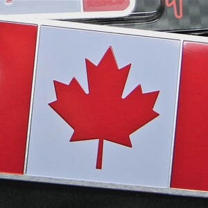 May include: Rectangular metal emblem featuring the Canadian flag. The design includes a red maple leaf on a white background, flanked by red vertical bars. The emblem has a silver border.