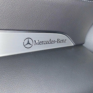Könnte beinhalten: Ein silberfarbenes Zierelement aus Metall mit dem schwarzen Mercedes-Benz Logo und Schriftzug. Die Leiste ist in eine dunkelgraue, texturierte Armaturenbrettoberfläche eingelassen und besticht durch ein klares Design mit gebürsteter Optik.