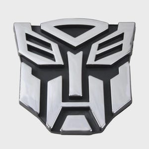 May include: A chrome-finished metal emblem featuring the Autobot insignia from the Transformers franchise. The design consists of a stylised robotic face with sharp, geometric lines and a polished silver surface set against a black background.