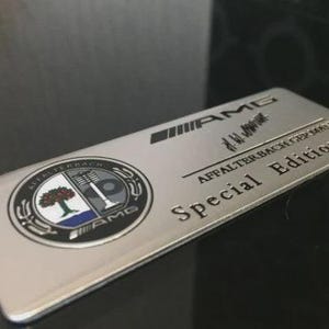 Puede incluir: Un emblema rectangular de metal plateado que presenta el logotipo de AMG, una firma y el texto Affalterbach Germany. Incluye un escudo circular con un gráfico de un árbol y una válvula, y las palabras Special Edition en relieve.