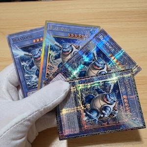 Puede incluir: Varias cartas coleccionables holográficas con el personaje Blastoise. Las cartas tienen una combinación de colores azul y dorado, con el personaje en varias poses de acción. El texto "BLASTOISE" es visible en cada carta.