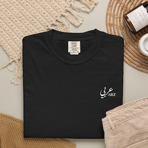 Puede incluir: Sudadera negra de cuello redondo con escritura árabe blanca y las letras "ARZ" bordadas en la parte delantera. La sudadera tiene una etiqueta que dice "Comfort Colors". La prenda está doblada sobre una superficie neutra.