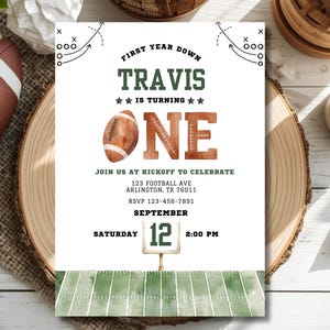 May include: A white birthday invitation card featuring a football theme with the text First Year Down Travis Is Turning One. The design includes a watercolor football, a green goal post with the number 12, and a green field at the bottom.