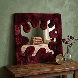 May include: A square mirror featuring a thick, tufted frame in varying shades of deep red and burgundy. The frame has an irregular, organic, wavy border that surrounds the central glass. The mirror reflects a stack of books and a patterned rug.