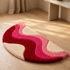 May include: A semi-circular rug featuring a tufted texture with wavy, flowing bands of color. The design consists of alternating stripes in deep red, vibrant hot pink, soft light pink, and cream, creating a bold, layered, and rhythmic pattern.