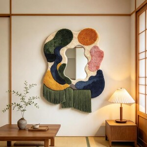 May include: An irregular wall mirror framed by a tufted textile border featuring abstract shapes in cream, dark green, navy blue, mustard yellow, orange, and pink, with long olive green fringe hanging from the bottom edge.