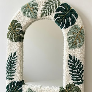 May include: An arched mirror with a white, textured frame adorned with green and light green leaf designs. The leaves are embroidered on the frame, creating a tropical, botanical theme. The mirror has a clean, modern aesthetic.