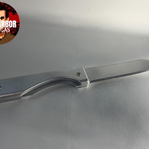 May include: A silver folding knife with a curved blade. The knife has a metallic handle and a visible hinge. The image includes a circular logo with the text "BAY HARBOR REPLICAS".