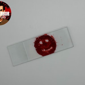 Puede incluir: Un portaobjetos de vidrio rectangular con una mancha roja circular en el centro. La sustancia roja está dispuesta para formar un patrón de cara sonriente simple con dos ojos y una boca curva visible dentro del pigmento carmesí.