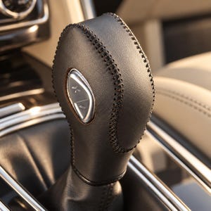 May include: A dark brown leather gear shift knob featuring prominent dark stitching along the edges and a silver-toned metallic button on the side. The gear shifter has a smooth, rounded top and a tapered base, integrated into a vehicle interior.