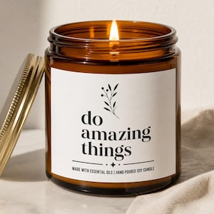 May include: A lit soy candle in an amber glass jar with a gold lid. The white label features a black botanical illustration, the text "do amazing things," and the phrase "MADE WITH ESSENTIAL OILS | HAND POURED SOY CANDLE" in black lettering.