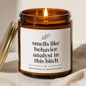 May include: A lit soy candle in a brown glass jar with a gold lid. The label reads "smells like behavior analyst in this bitch." The candle is hand-poured and made with essential oils.