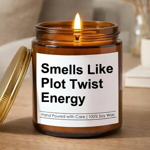 May include: A lit soy wax candle in a brown glass jar with a gold lid. The label reads "Smells Like Plot Twist Energy." The candle is hand poured with care.