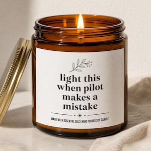 May include: A lit soy candle in a brown glass jar with a gold lid. The label reads "light this when pilot makes a mistake." The candle is hand-poured and made with essential oils.