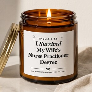 May include: A lit brown glass jar candle with a gold lid. The candle has a white label with the text "Smells Like I Survived My Wife's Nurse Practitioner Degree". The candle is made with essential oils and hand-poured soy.