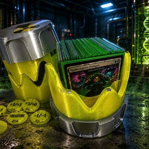 May include: A yellow and silver radioactive waste barrel-shaped card deck box containing a stack of trading cards, with the top card titled Humongous Fungus. Several yellow circular tokens with markings like +1/+1, +1/+0, -1/-1, and Treasure are scattered nearby.