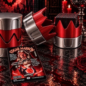 May include: A red and silver cylindrical card deck box with a crown-shaped lid, featuring a metallic finish. Beside it lies a collectible trading card depicting a vampire character named Edgar Markov with text detailing game abilities and stats.