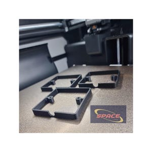 May include: Four black, square-shaped 3D-printed components with rounded corners and mounting holes are arranged on a light brown surface. The components are in front of a 3D printer. A logo that says "SPACE TECH" is visible.
