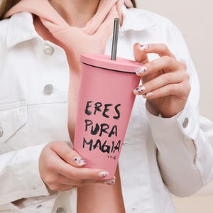 Puede incluir: Un vaso rosa con tapa negra y pajita plateada. El vaso tiene la frase "ERES PURA MAGIA" impresa en negro. El vaso es sostenido por una persona que lleva una chaqueta blanca.