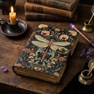 Peut inclure: Un journal rectangulaire avec une couverture sombre ornée d'une grande libellule dorée et d'un motif floral. Le livre possède une fermeture à loquet en métal et des pages en papier vieilli aux bords irréguliers avec des illustrations botaniques.