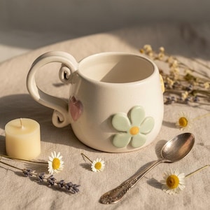 Peut inclure: Une tasse en céramique couleur crème avec un corps arrondi, une anse décorative enroulée, un cœur rose en relief et une grande fleur vert clair au centre jaune. La tasse est accompagnée d'une petite bougie, d'une cuillère en argent et de fleurs.