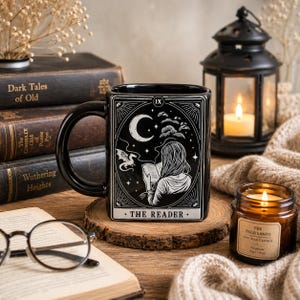 Peut inclure: Un mug en céramique noire orné d'une illustration graphique blanche représentant une personne lisant un livre sous un croissant de lune et des étoiles, avec un petit dragon. Le motif est encadré comme une carte de tarot marquée IX THE READER.