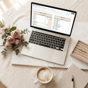 May include: A laptop displaying a wedding planning spreadsheet sits on a marble surface, surrounded by a bouquet of pink roses, a cup of coffee, a notebook, and a ring box. The screen shows a budget table and timeline.
