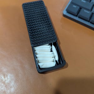 May include: A rectangular black plastic sliding case with a textured, wavy pattern on the lid. The case is partially open, revealing a row of small, rectangular, off-white pouches neatly arranged inside the storage compartment.
