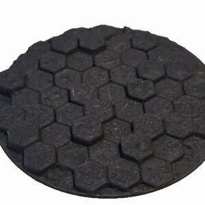 May include: A round, dark gray object with a textured surface. The surface is covered in a pattern of hexagonal shapes. The object appears to be made of a composite material.